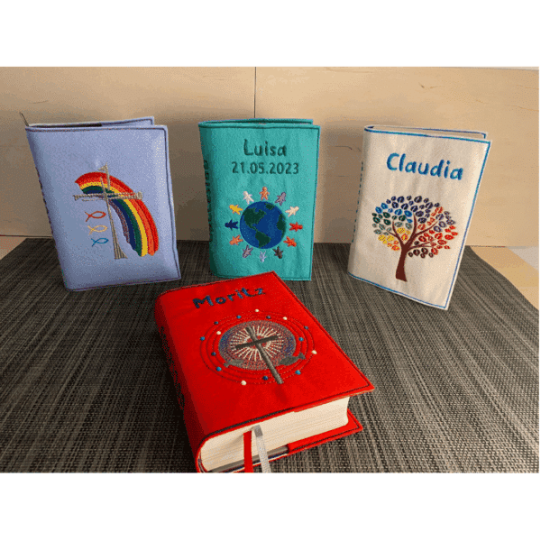 Vier bunte, individuell gestaltete Gotteslobhüllen aus Filz, jede mit einem einzigartigen Motiv, darunter ein Kreuz mit Regenbogen, eine Weltkugel, ein bunter Baum und eine Pusteblume. Die Buchhüllen tragen Namen und Daten.
