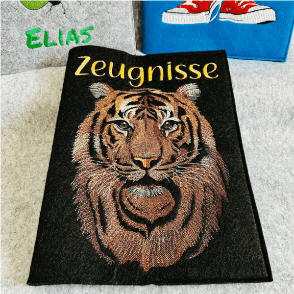 Personalisierte Zeugnismappe aus Filz mit gesticktem Tigerkopf und dem Wort "Zeugnisse" darüber. Im Hintergrund grauer und blauer Stoff mit bunten Verzierungen.