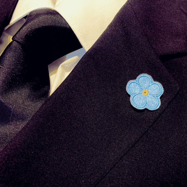 Forget Me Not Anstecker in einfarbig hellblau am Revers eines schwarzen Sakkos, getragen.