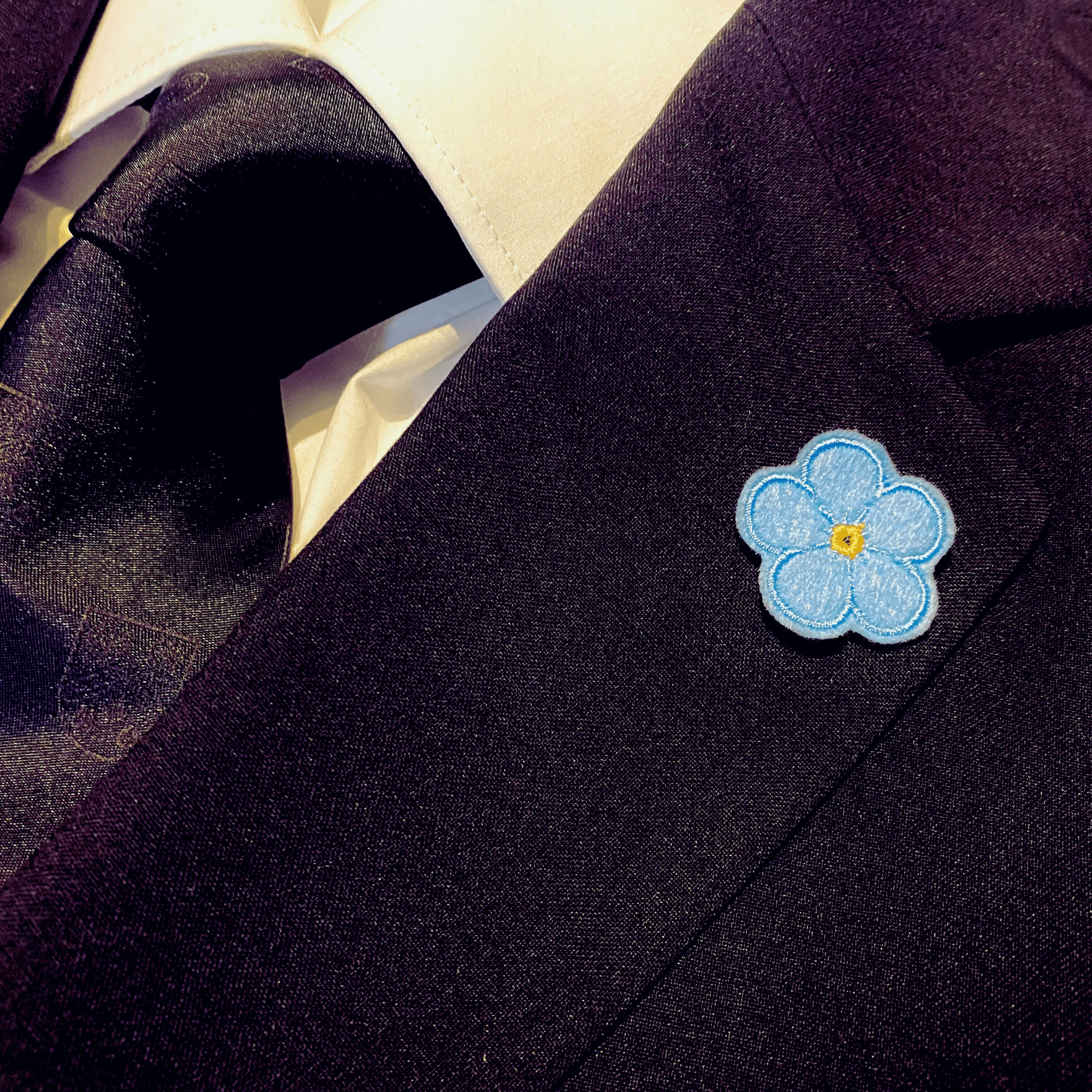 Forget Me Not Anstecker in einfarbig hellblau am Revers eines schwarzen Sakkos, getragen.