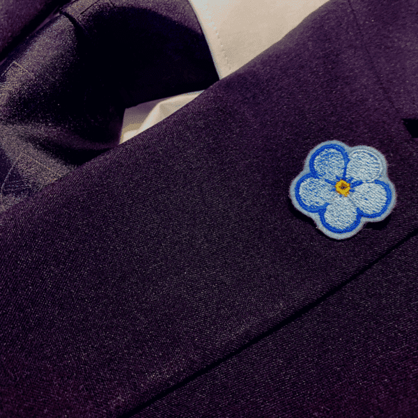 Forget Me Not Anstecker mit mehrfarbig blauer Kontur auf dem Revers eines Sakkos, getragen.