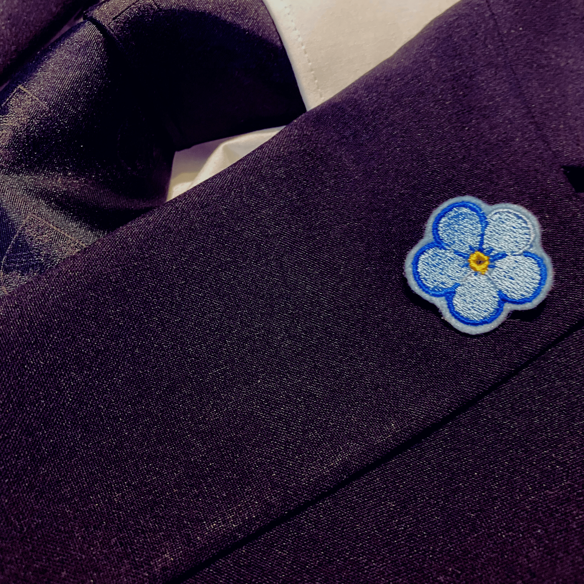 Forget Me Not Anstecker mit mehrfarbig blauer Kontur auf dem Revers eines Sakkos, getragen.