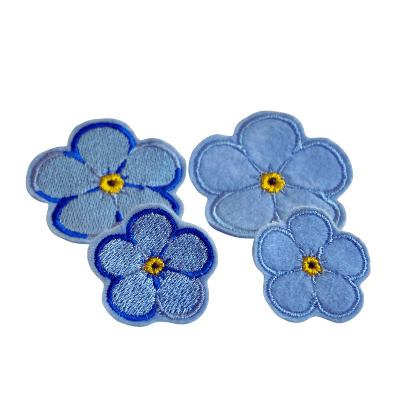 Forget Me Not Anstecker in vier Varianten als gestickte blaue Blüten mit gelber Mitte, Nahaufnahme.