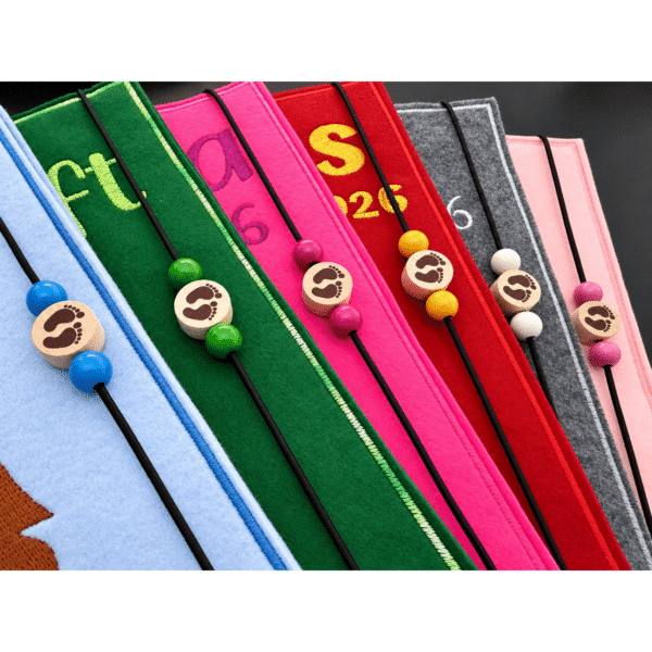Detailaufnahme des Gummiband-Verschlusses einer U-Heft-Hülle aus Filz mit zwei farbigen Holzperlen und Motivperle mit Babyfüßen
